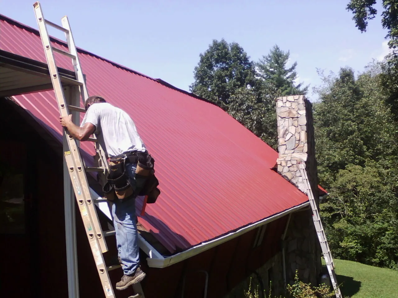 Roofer on a ladder beside a red metal roof and stone chimney — Soffit & Fascia Repair by Roofing Brothers in Shandaken